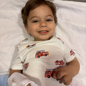 Toddler with dark hair and light skin wearing brace on right arm lying in hospital bed smiling.