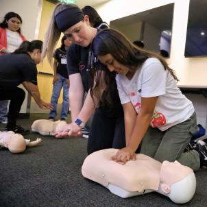 Two women with a light skin tone wearing uniforms and kneeling down to gesture to mannequins on the floor, on which high school-age kids are practicing CPR.