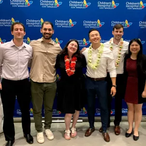 Four young men and two young women dressed in business casual attire stand arm in arm in front of blue Children's Hospital Los Angeles background, flanked by two pillars of white balloons