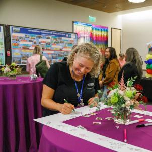 A woman stands at a table signing her name on a commemorative paper picture frame. 