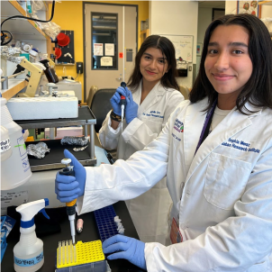 Two high school girls in lab coats smile at the camera while standing in a scientific laboratory.