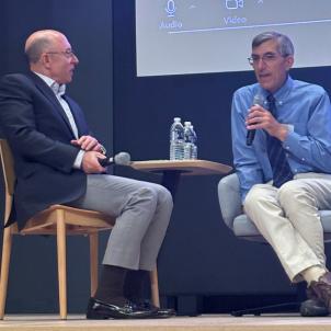 A man in a navy suit jacket sits next to a gray-haired man who is talking into a microphone and wears a blue shirt, tie and light pants.