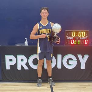 High school-aged Hunter stands holding a basketball trophy wearing a blue and yellow "PRODIGY" basketball uniform. 