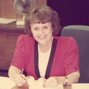 A woman with light skin tone, short dark hair wearing a red dress sitting on a chair in an office.