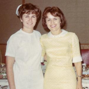 An interior picture of a light skin tone young woman in a nurse outfit with short dark hair standing next to a woman with short dark hair in a yellow dress.