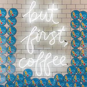 Lit up marquee sign with words but first, coffee surrounded with circular cards of colorful butterflies against a beige brick wall