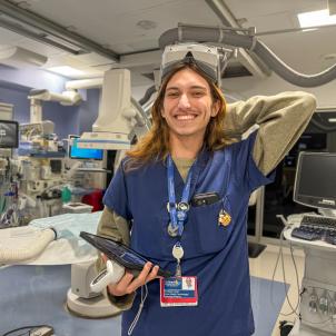 Phoenix Hunt stands smiling in the Interventional Radiology procedure room wearing VR goggles on his forehead and holding an iPad. 