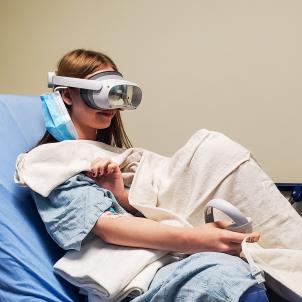 A young girl sits on a hospital bed wearing VR goggles and holding a VR remote.  