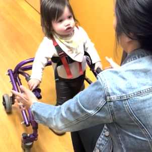 A toddler walks toward her physical therapist using a walker. 