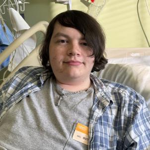 A young man in a hospital bed and dressed in a T-shirt and plaid shirt looks out at the camera with a peaceful expression.