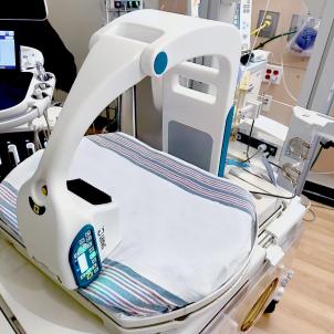 A white curved machine in the shape of a C is shown over a baby's isolette bed. A computer monitor and tubing and equipment are in the background.