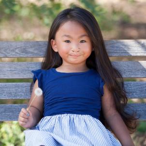 A young girl with medium skin tone and long, dark hair wearing a blue shirt and striped skirt sitting on a bench outside.