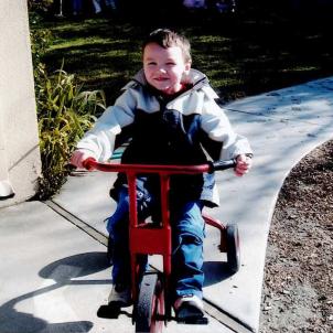 Young Ryan rides a red tricycle outdoors.