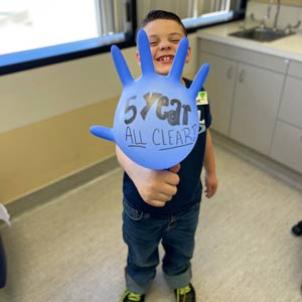 Young boy with light skin tone smiles as he holds a blown up surgical glove with the text "5 year all clear!"