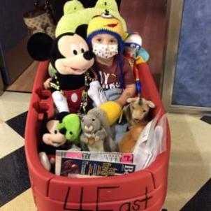 Young boy with light skin tone wearing a yellow beanie and surgical mask surrounded by stuffed animals