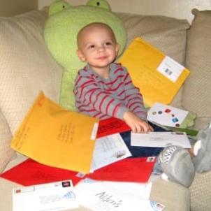 Young boy with light skin tone and shaved head sits on couch surrounded by envelopes