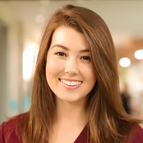 Nurse with light skin tone and auburn hair wearing a maroon nurses uniform and holding a binder smiles as she stands in a hospital corridor