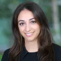 Headshot of woman with light skin tone and dark hair wearing black suit jacket with blurred outdoor background