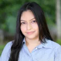 Headshot of a woman with medium skin tone and dark hair wearing a blue blouse with a blurred outdoor background