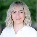 Headshot of woman with light skin tone and light hair wearing white blouse with blurred outdoor background