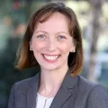 Headshot of a smiling woman with light skin tone and short auburn hair wearing a white top under a grey suit jacket against a blurred outdoor background