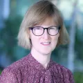 Headshot of a smiling woman with light skin tone and short blonde hair wearing dark-rimmed glasses and a purple patterned top against a blurred outdoor background