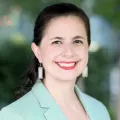 Headshot of a smiling woman with light skin tone and dark hair wearing a mint green suit jacket against a blurred outdoor background