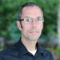 Headshot of a man with light skin and short grey hair wearing glasses and a black buttoned-tone shirt against a blurred outdoor background