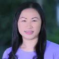 Headshot of a smiling young woman with light skin tone and straight dark hair wearing a purple top against a blurred outdoor background