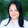 Headshot of smiling woman with light skin tone and dark hair wearing lavender top against a blurred outdoor background