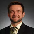 Headshot of a smiling man with light skin tone, brown hair and a light beard wearing a yellow dress shirt, blue and green striped tie and dark suit jacket against a dark neutral indoor background