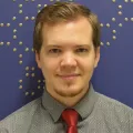 Headshot of smiling young man with light skin tone and brown hair wearing a grey dress shirt and red tie against a blue indoor background