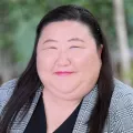 Headshot of a smiling woman with light skin tone and dark hair wearing a black blouse under a grey plaid patterned suit jacket against a blurred outdoor background