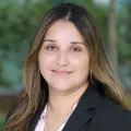 Headshot of a smiling woman with medium skin tone and long brown hair wearing a light pink blouse under a dark suit jacket against a blurred outdoor background