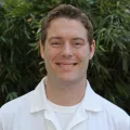 Headshot of a smiling man with light skin tone and short brown hair wearing a white t-shirt under a white lab coat against a blurred outdoor background