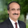 Headshot of a smiling man with medium skin tone, moustache and receding dark hair wearing a white dress shirt and maroon checkered tie under a dark suit jacket against a blurred outdoor background