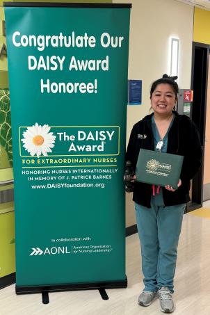 Woman with medium skin tone and pulled-back dark hair wearing blue scrubs under a dark fleece jacket smiles as she poses with her DAISY Award next to a standing DAISY Award poster in a hospital corridor