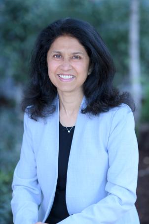 Headshot of a smiling woman with medium skin tone and shoulder-length dark hair wearing a black top under a light blue suit jacket against a blurred outdoor background