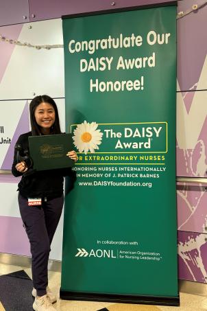 A young woman with medium skin tone and straight dark hair smiles as she poses with her DAISY Award statue next to a DAISY Award poster