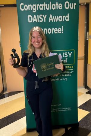 Woman with light skin tone and blonde hair wearing dark colored nurses uniform smiles as she poses with her DAISY Award statute