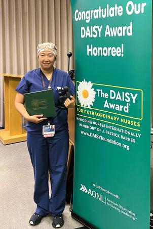 Female nurse with medium skin tone and wearing blue scrubs smiles as they pose with their DAISY Award next to a DAISY Award poster