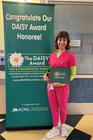 A female nurse with light skin tone and short brown hair smiles as she poses with her DAISY Award next to a poster that reads "Congratulate Our DAISY Award Honoree!"