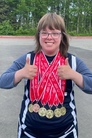 Medium shot of a light-skinned young adult woman standing outside and giving two thumbs up, wearing athletic attire and several Special Olympics medals