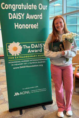 Female nurse with light skin tone and blonde hair smiles as they pose with their DAISY Award next to a DAISY Award poster