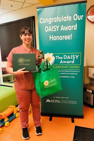 Smiling female nurse with light skin tone and dark hair poses with their DAISY Award next to a DAISY Award honoree poster