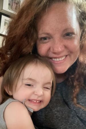 Mother with light skin tone and auburn hair smiles as she gets a hug from her young daughter
