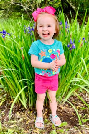 Toddler with light skin tone wearing bright pink bow and colorful t-shirt smiles as she stands next to purple daffodils growing in the ground