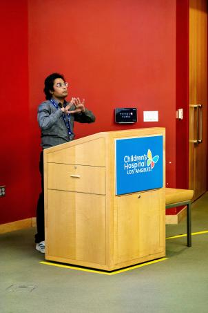 A young adult man with medium-dark skin tone stands at a lectern with the Children’s Hospital Los Angeles logo on it in front of a red wall in an auditorium, gesturing as he delivers a presentation.