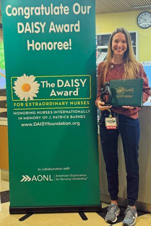 Female nurse with light skin tone and blonde hair smiles as they pose with their DAISY Award next to a DAISY Award poster