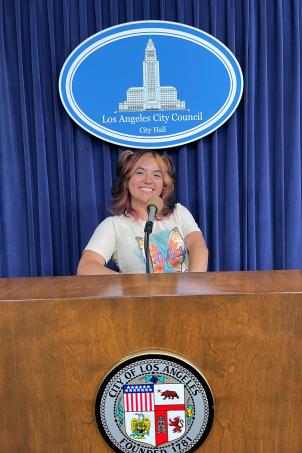 Medium shot of a high-school age kid with a medium skin tone and shoulder-length hair standing indoors behind a wooden lectern with the City of Los Angeles seal on it and a blue curtain with the Los Angeles City Council logo on it.